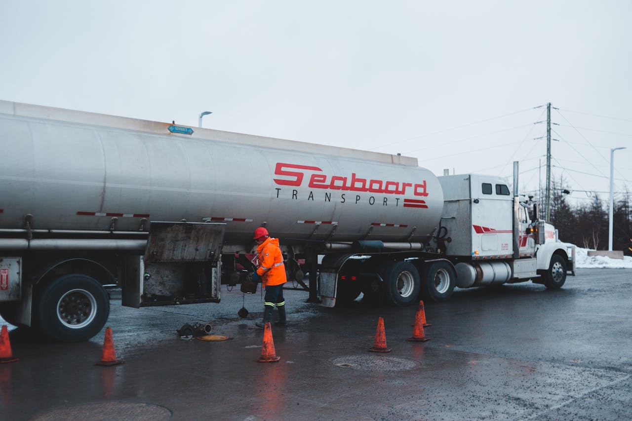 creative-02 Back view of anonymous male worker in bright uniform and helmet cleaning oil truck vehicle while standing on asphalt roadway surrounded by barriers under white sky in town